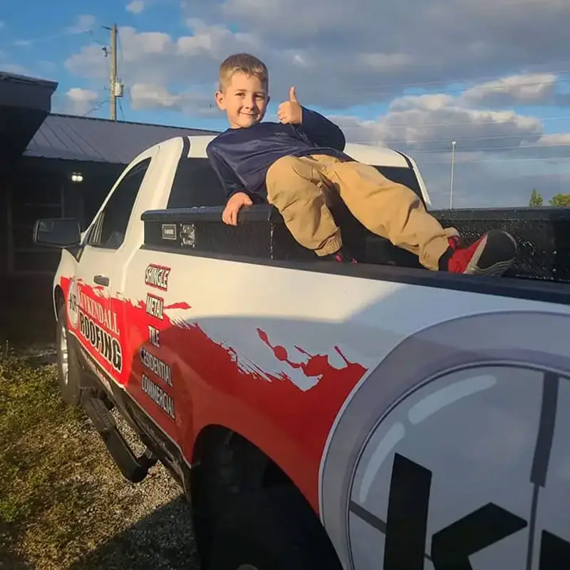 Smiling child giving a thumbs-up while sitting on a branded Kuykendall Roofing truck, representing family-owned values and community pride